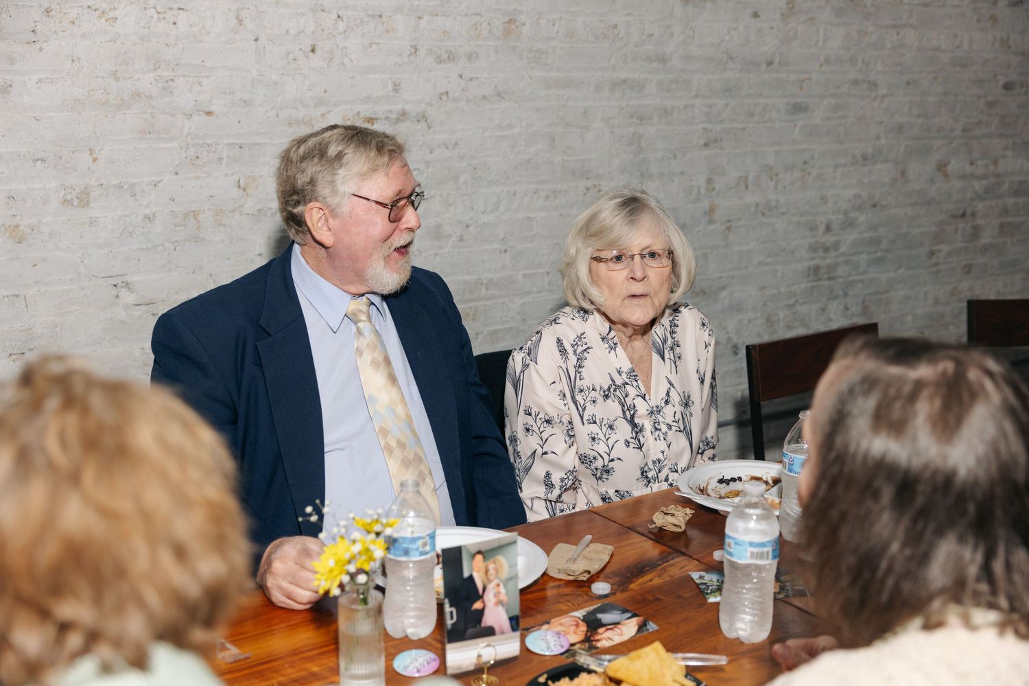Elderly couple seated together at wooden reception table with white brick wall background during wedding celebration.