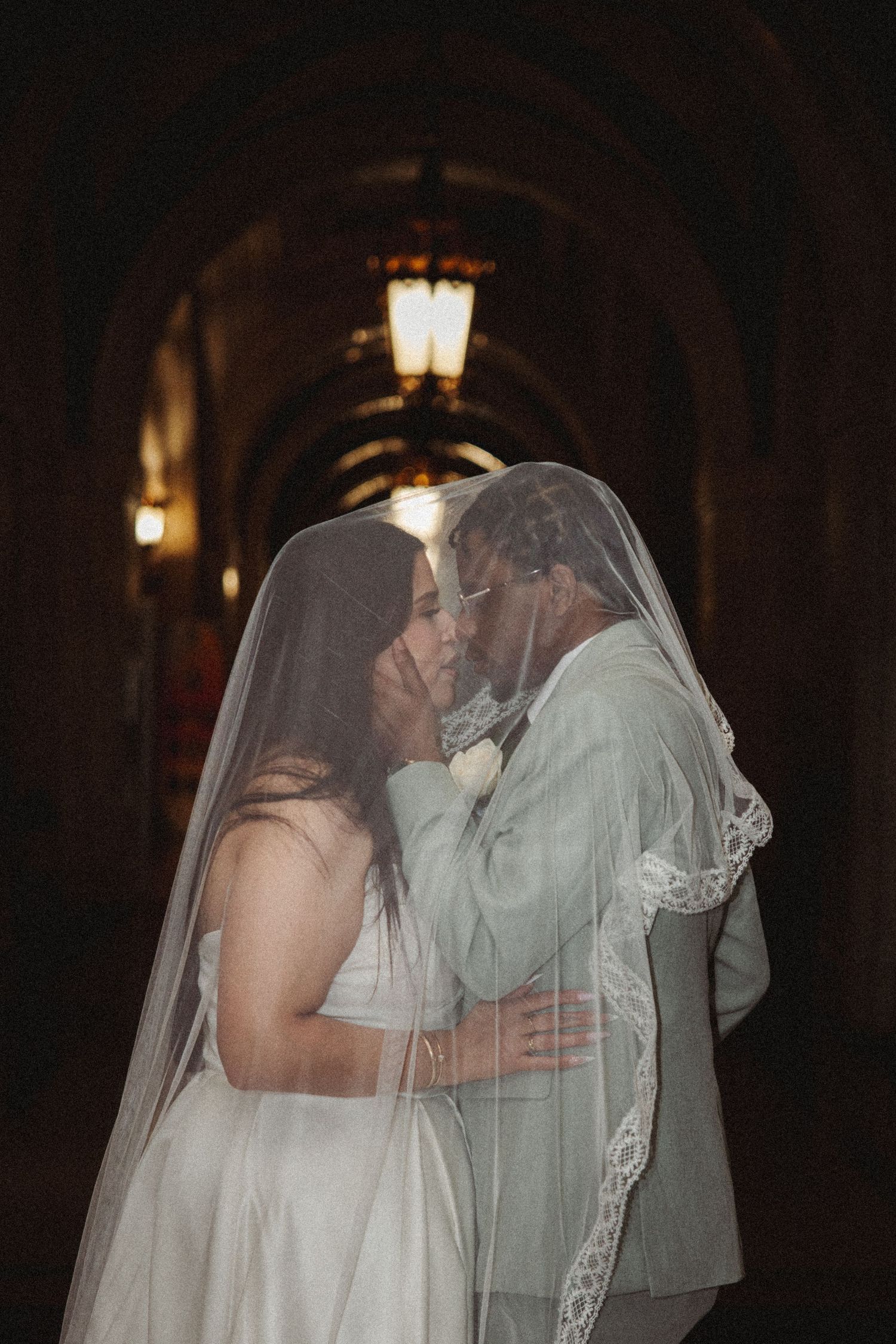 Bride and groom share a romantic kiss under a wedding veil in a dimly lit arched hallway.