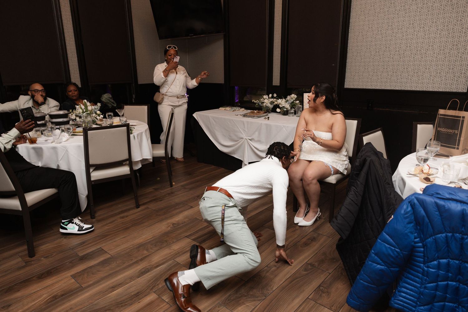 Wedding guests celebrate as groom kneels before bride during fun reception game at elegant venue.