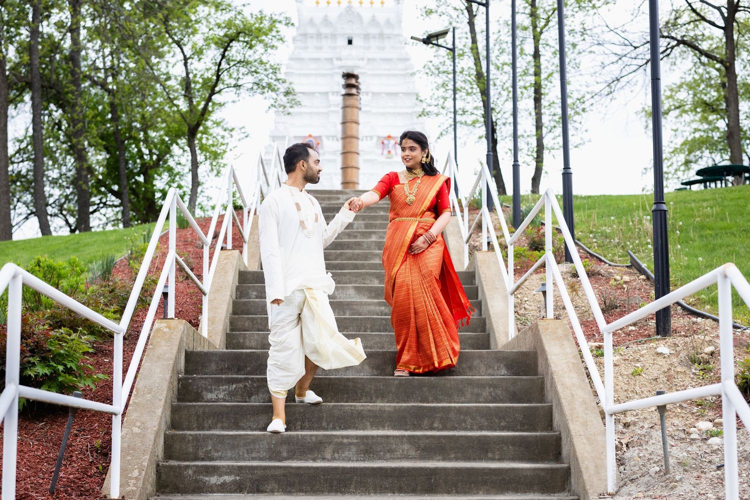Indian wedding couple holding hands walking down outdoor stone steps near white temple.