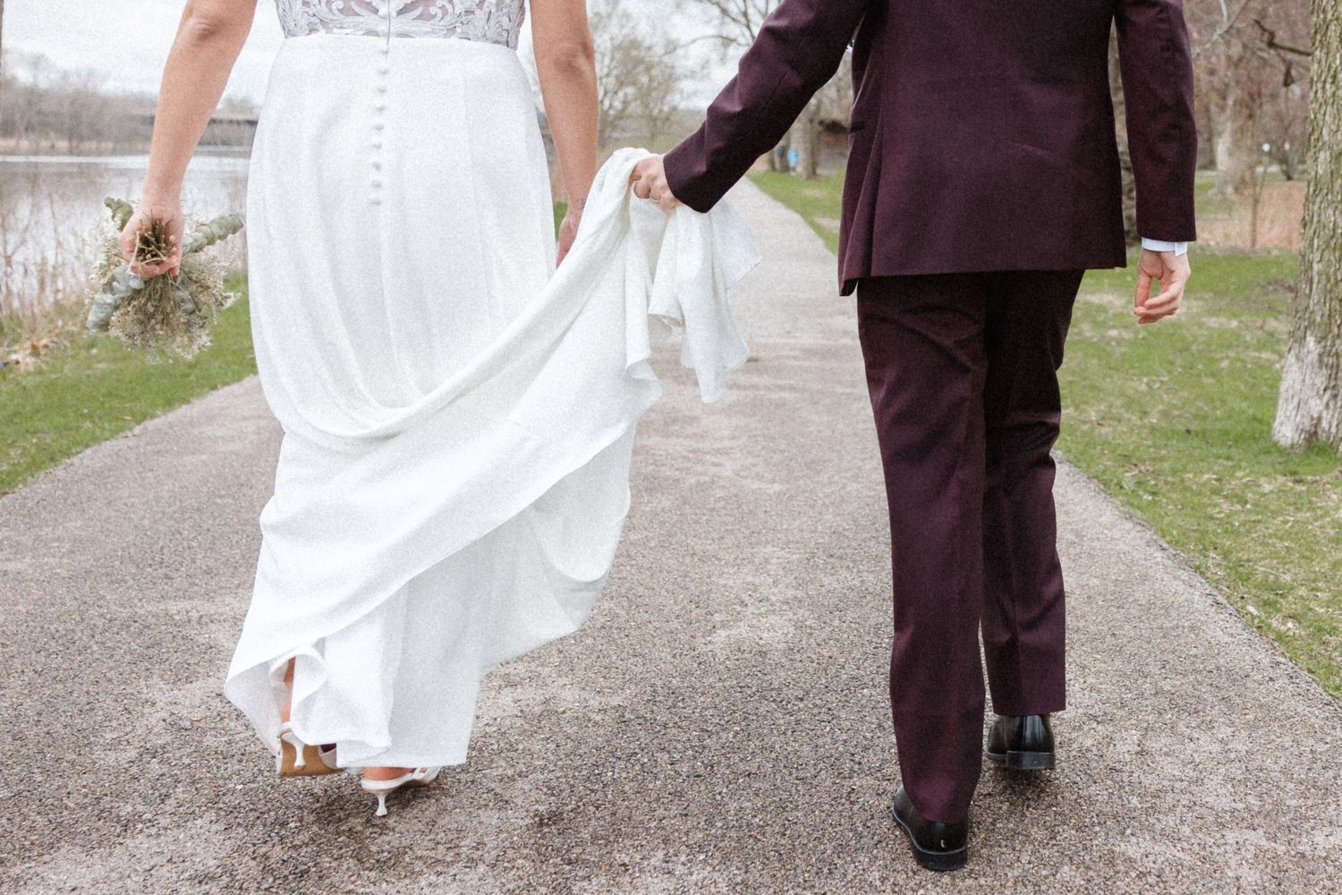 Bride and groom walking hand in hand on a path, bride in white gown, groom in burgundy suit.