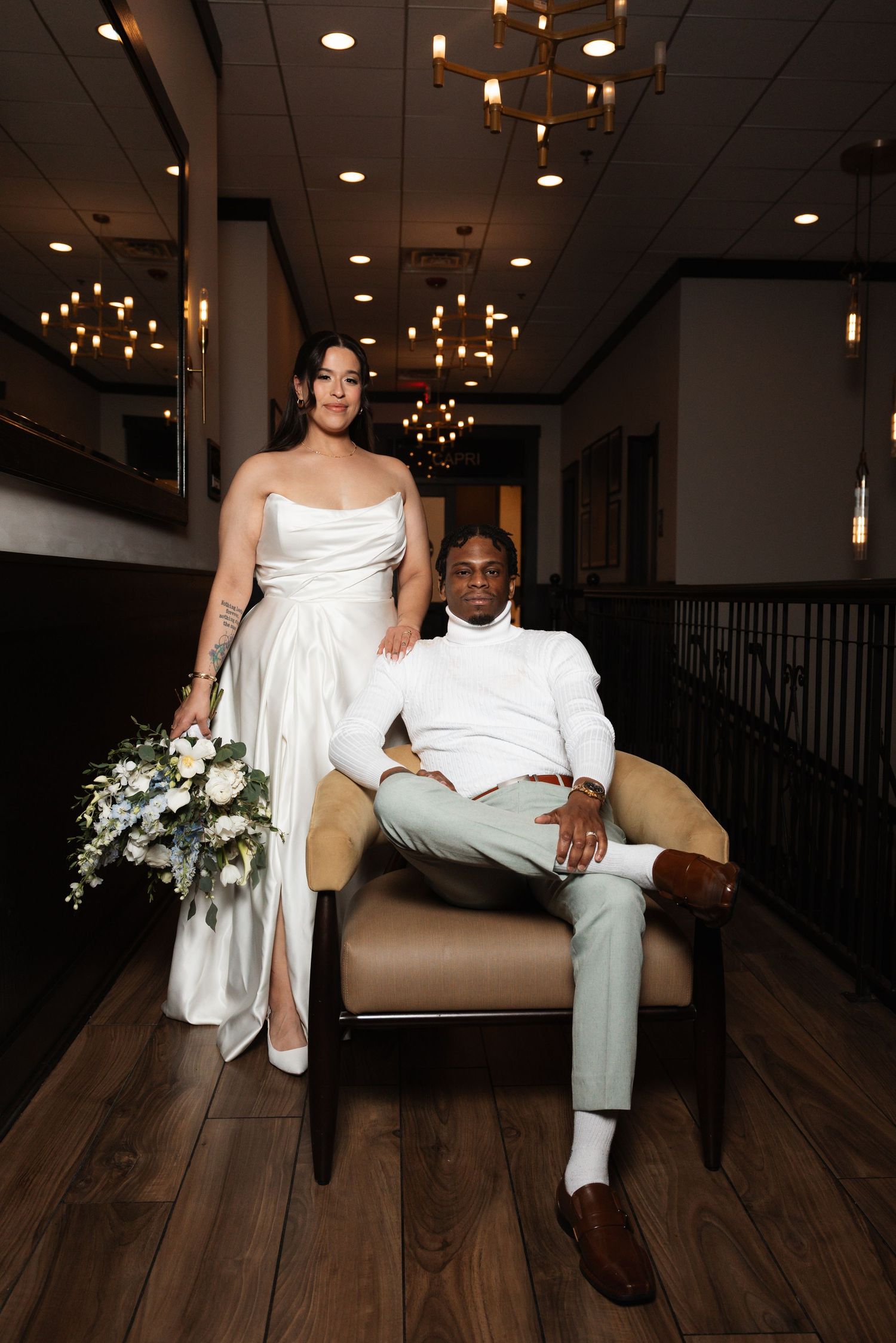 Bride with floral bouquet standing beside groom seated in leather chair under chandelier.
