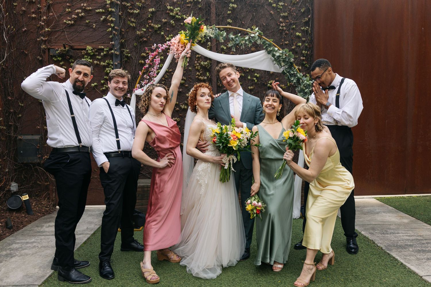Wedding party posing under floral arch, bridesmaids in colorful dresses with groomsmen in black suits.
