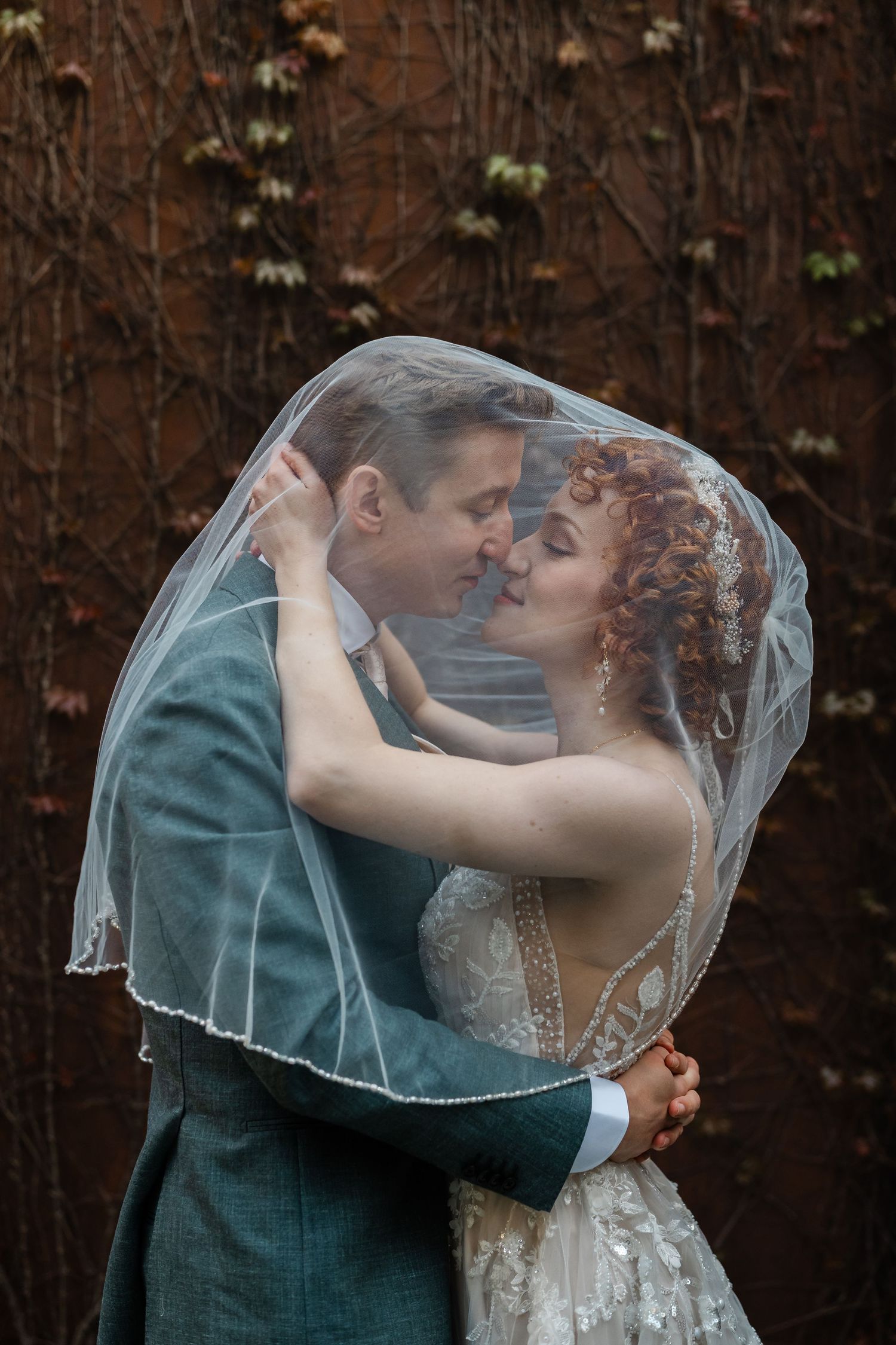 Bride and groom share romantic moment under sheer wedding veil, nearly kissing against rustic vine-covered backdrop.