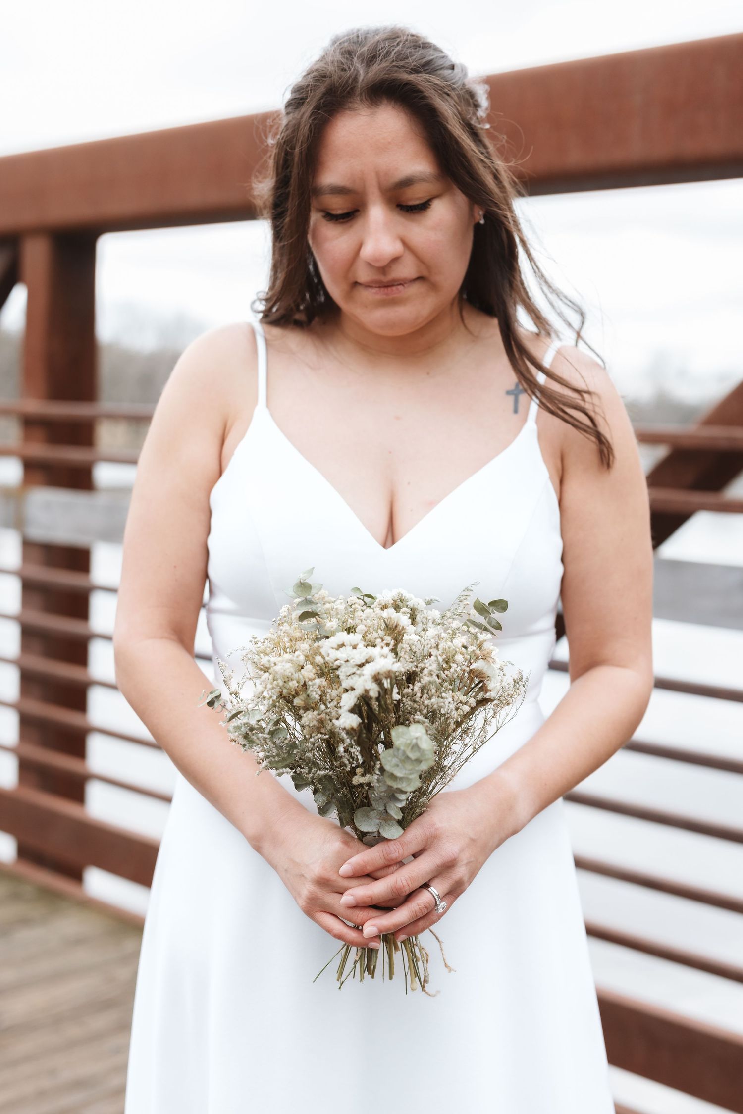 Bride in white spaghetti strap dress holds dried wildflower bouquet on wooden bridge, eyes downcast.