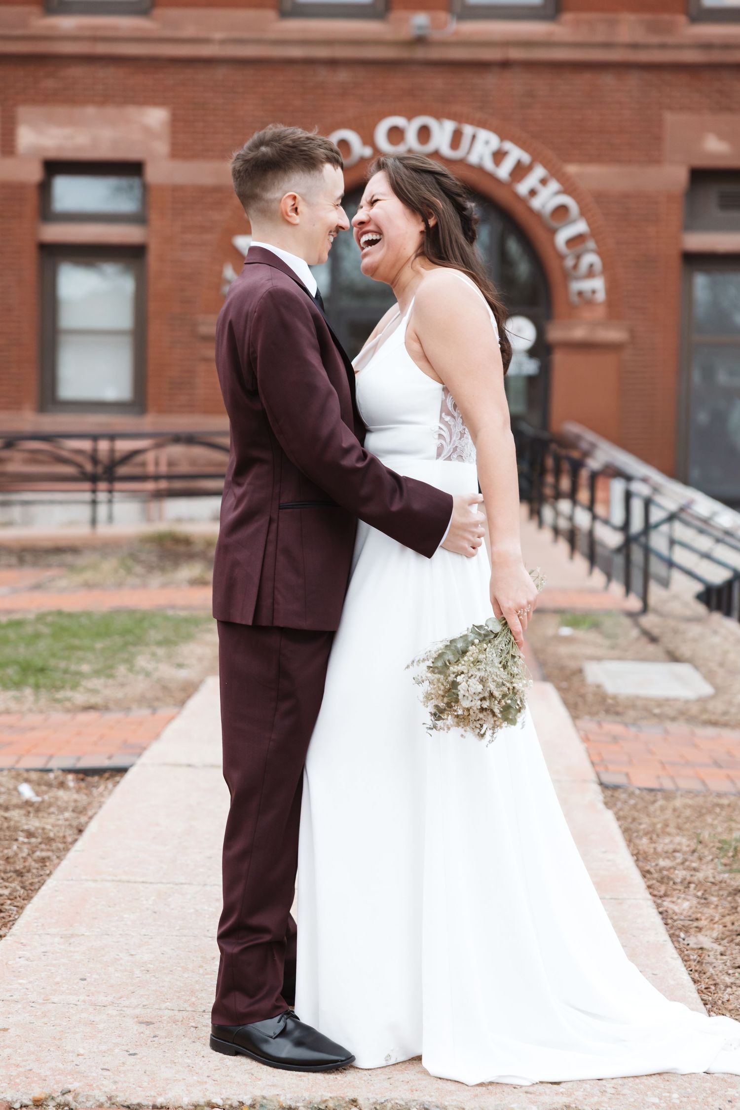 Bride and groom laughing together outside a brick courthouse, she holds a bouquet in a white gown, he wears a burgundy suit.