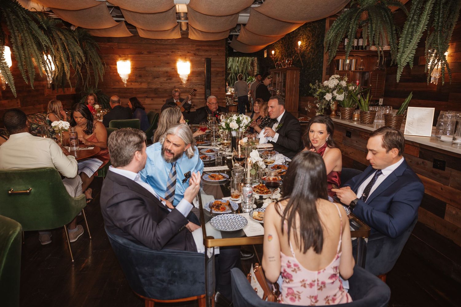 Wedding reception dinner guests socializing at elegantly decorated tables in warm lit venue.