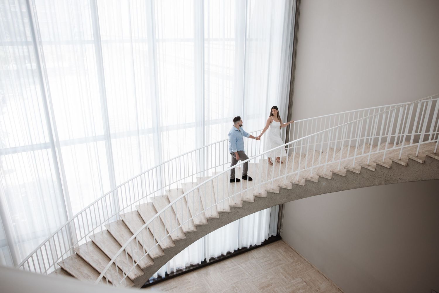 Couple holding hands walking down elegant curved white staircase in modern building with floor-to-ceiling windows.
