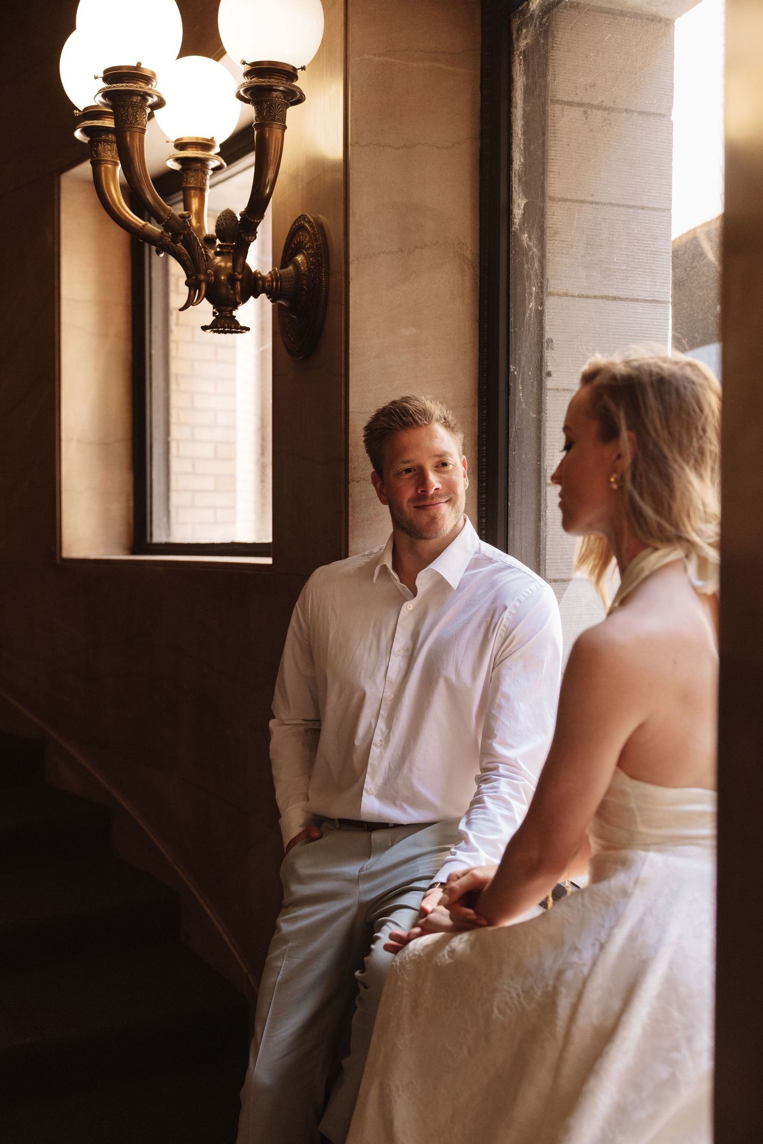 Groom in white shirt holds bride's hand while smiling at her near an ornate brass wall sconce.