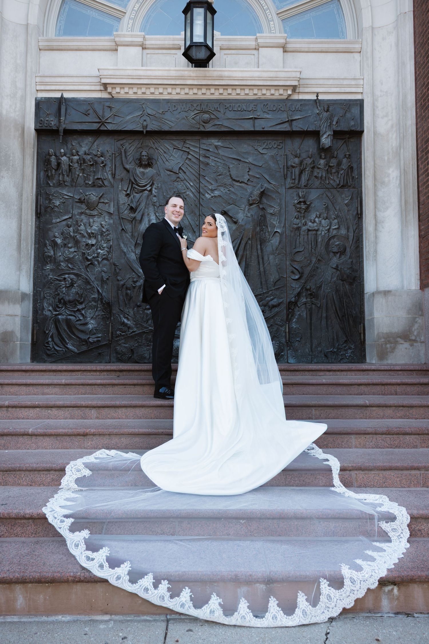 Bride and groom pose on church steps before ornate bronze doors, long lace veil trailing.