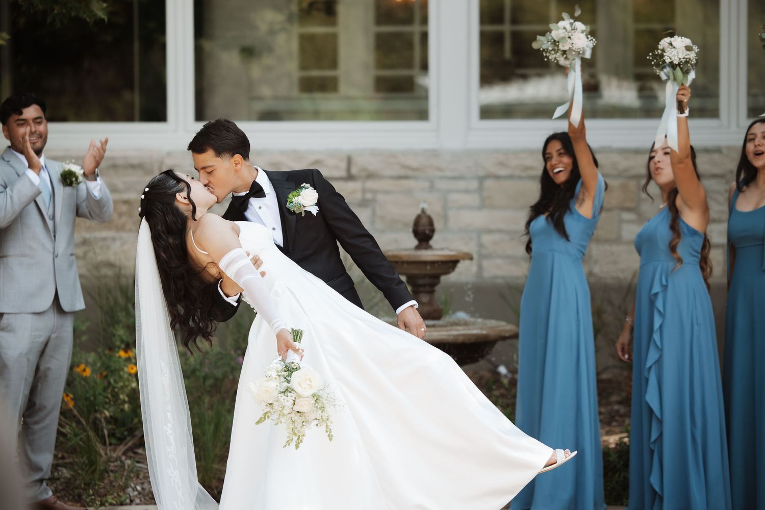 Groom dramatically dips bride while bridesmaids in blue dresses cheer and raise bouquets outdoors.