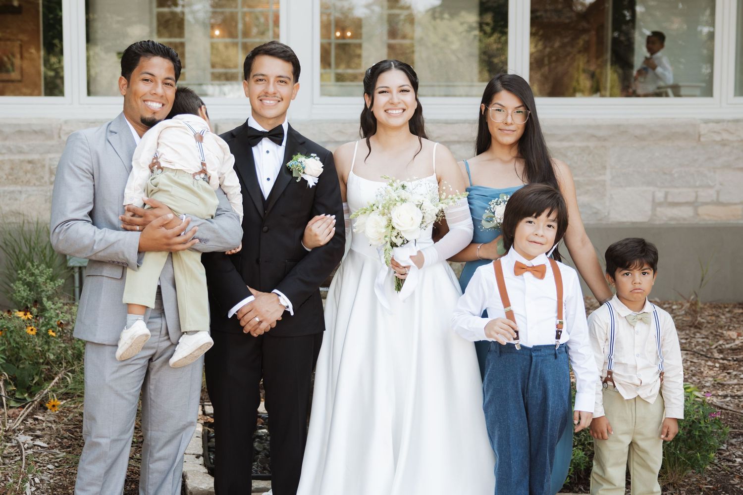 Bride and groom smile with family group including children outside elegant wedding venue.