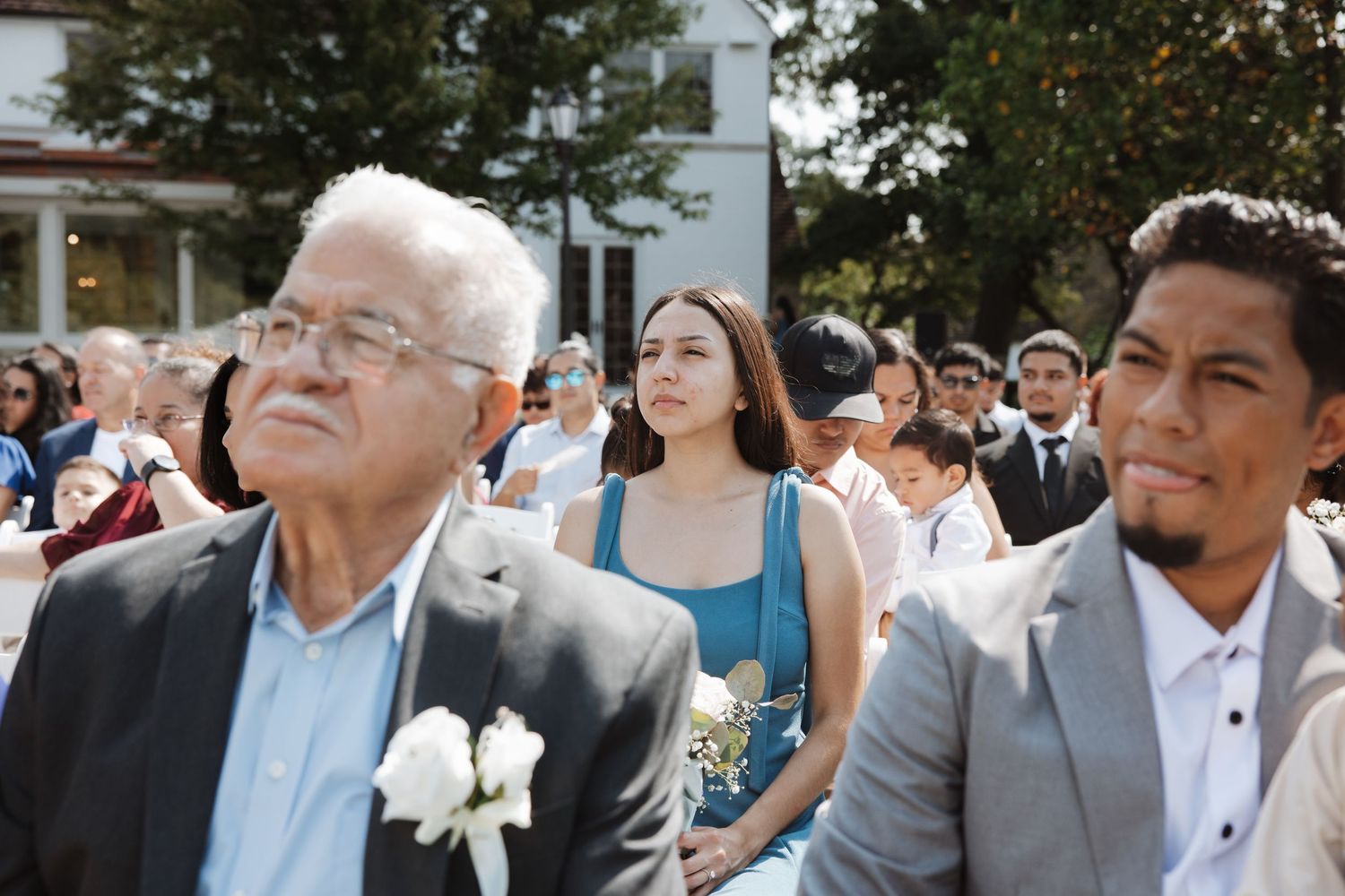 An elderly man and younger guest watch attentively during an outdoor wedding ceremony on a sunny day.