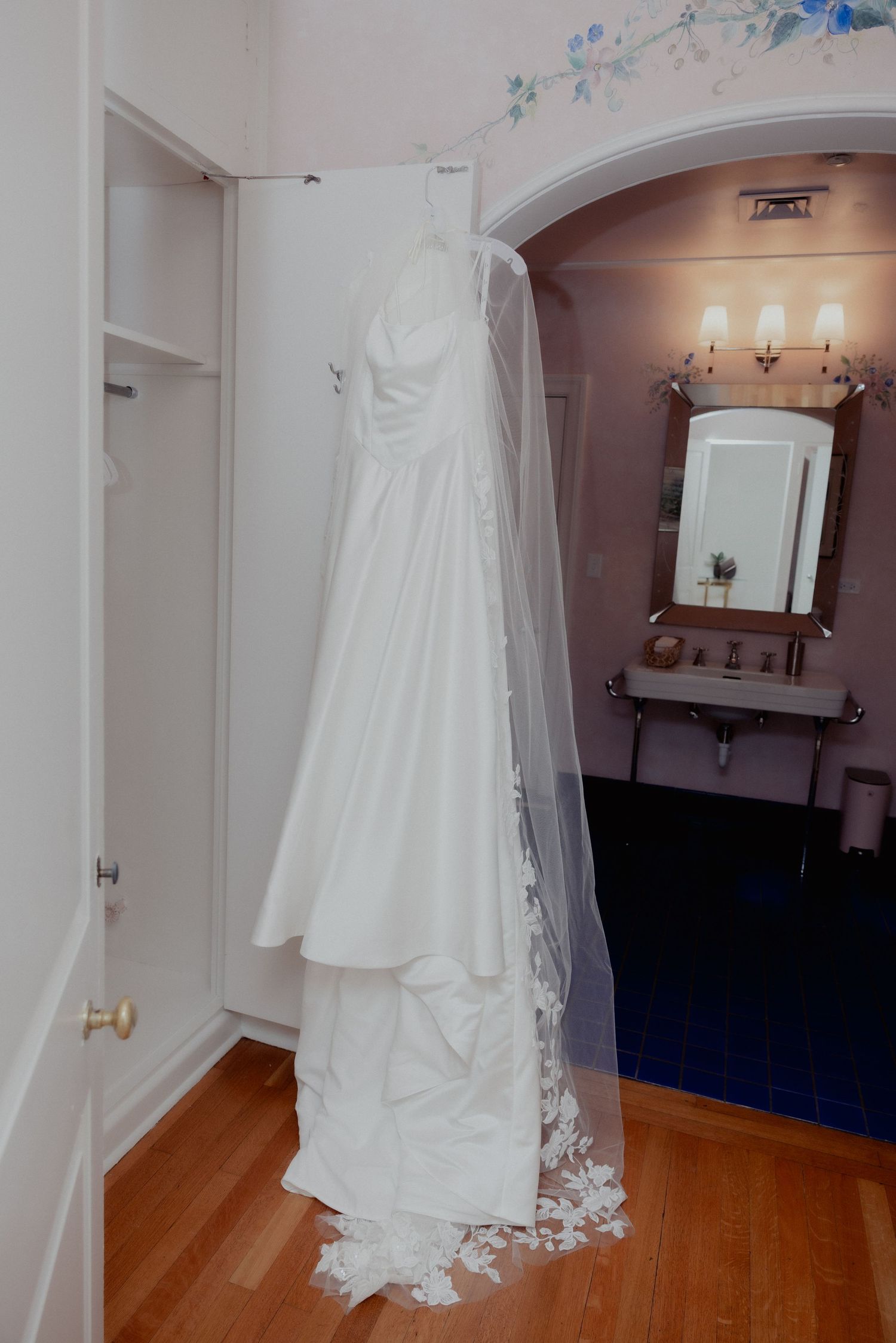 White wedding dress with veil hanging on closet door near elegant bathroom with mirror and wall sconces.