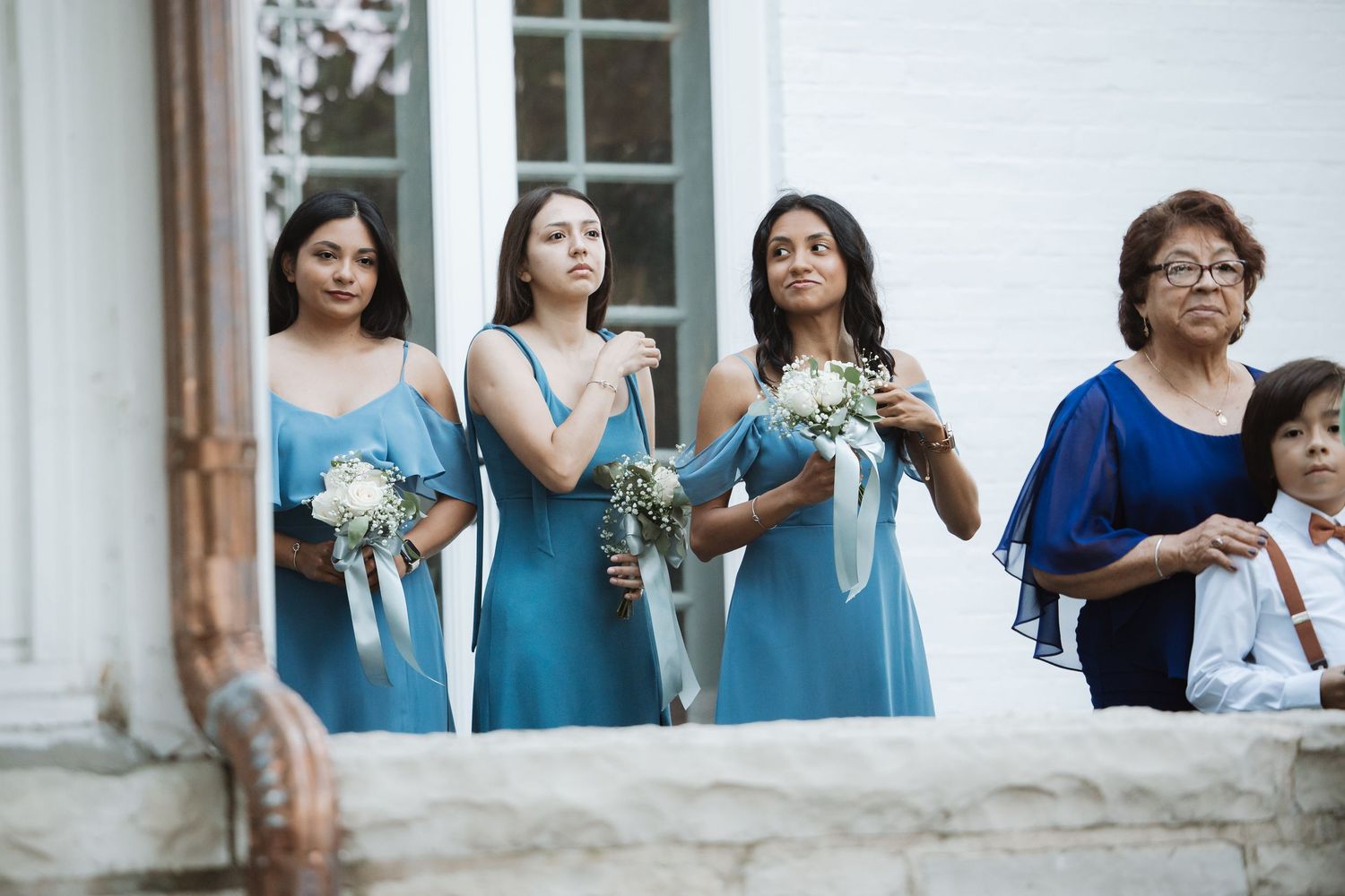 Three bridesmaids in teal off-shoulder dresses holding white bouquets at outdoor wedding ceremony.