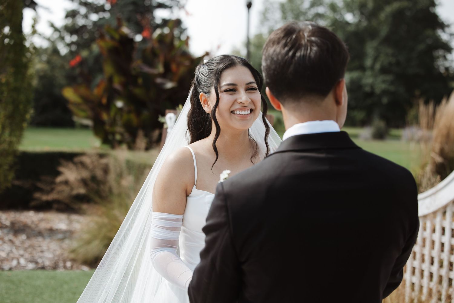 Radiant bride smiling at groom during outdoor wedding ceremony in lush garden setting.