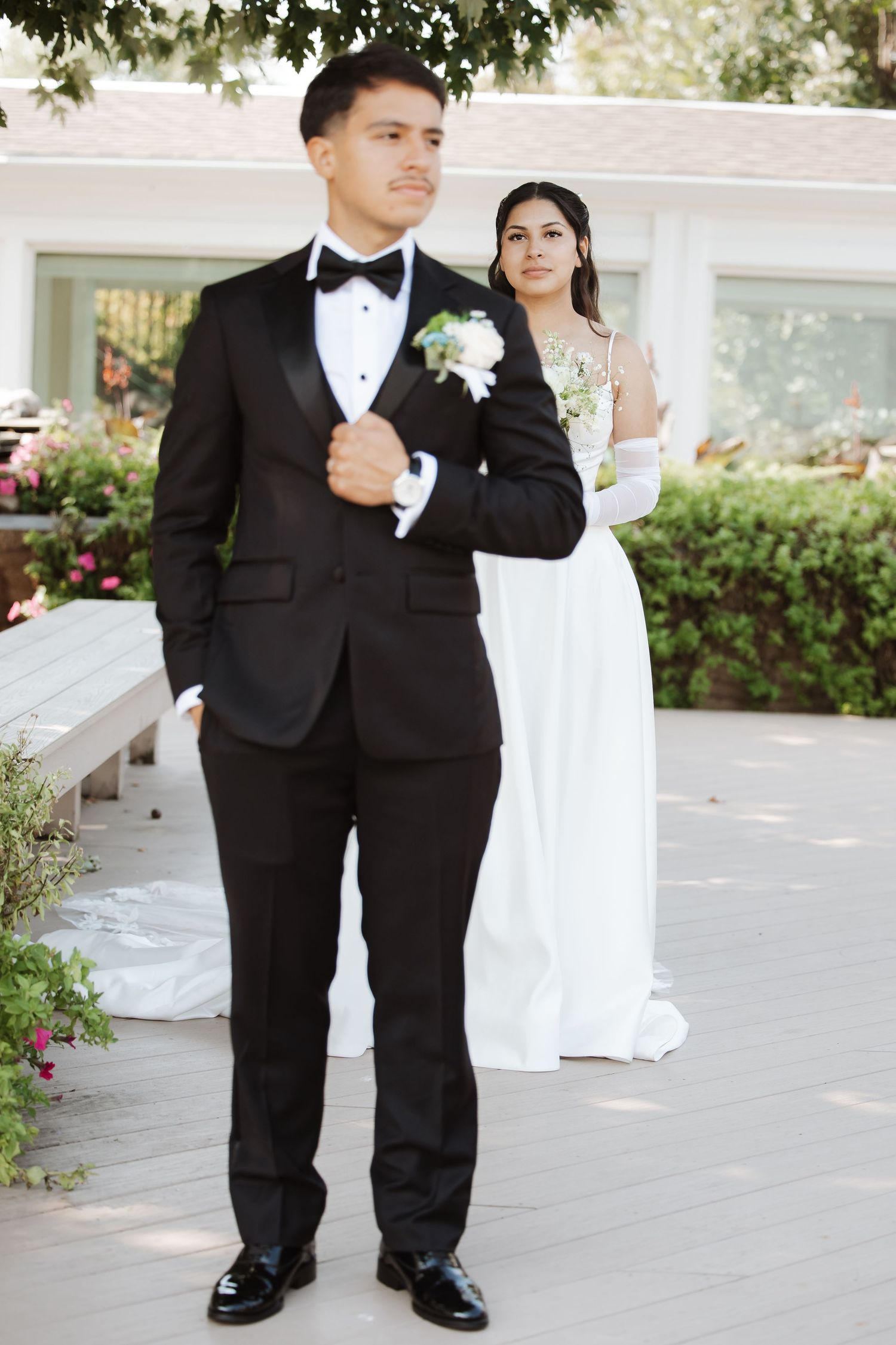 Bride in white gown stands behind groom in black tuxedo adjusting jacket lapel outdoors.