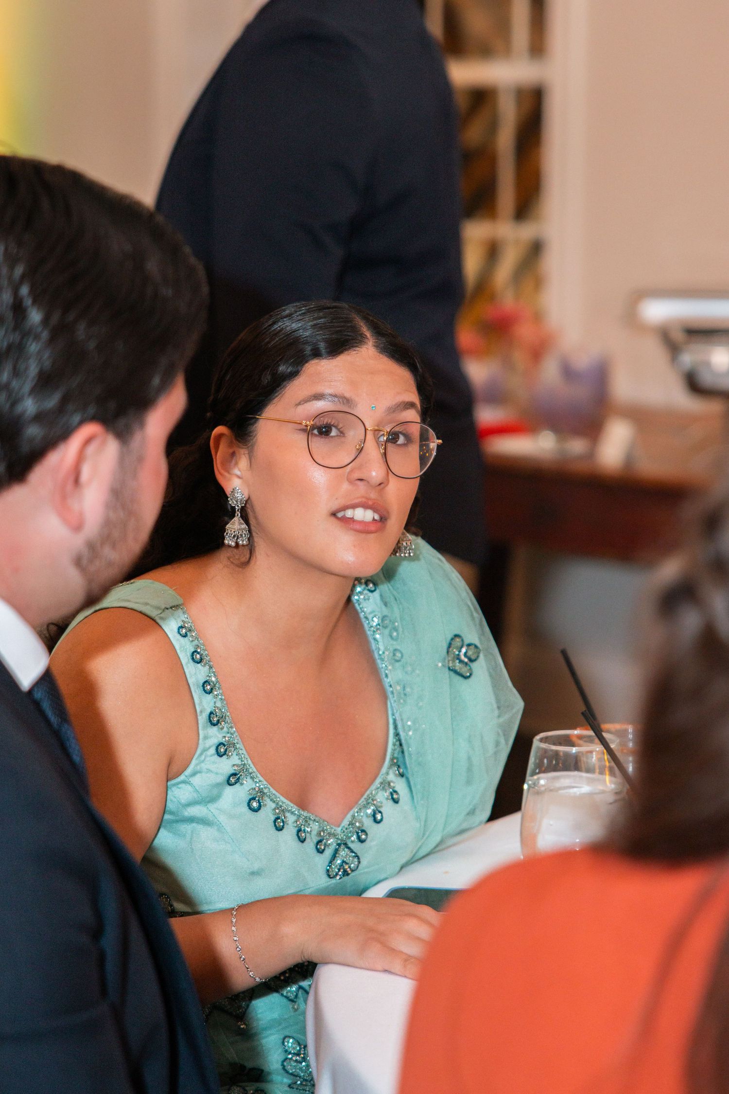 Elegant woman in teal embroidered saree with glasses engaged in conversation at an Indian wedding reception.