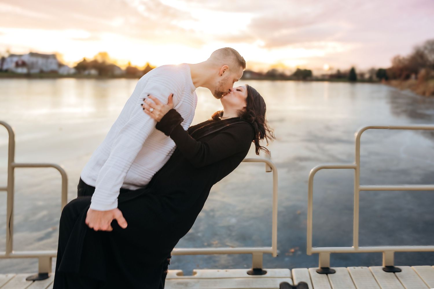 Romantic couple sharing a dip kiss at sunset on a waterfront dock, dressed in black and white outfits.