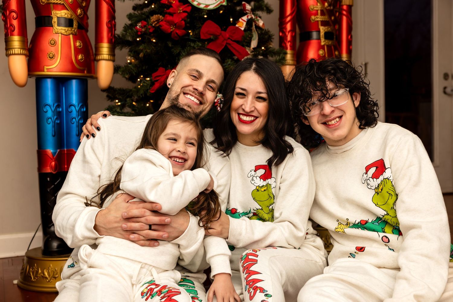 Family of four in Grinch holiday pajamas sitting together by Christmas tree.