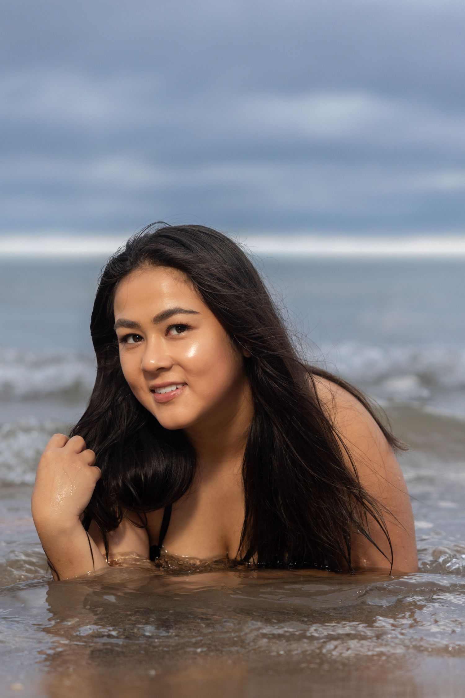 Young woman with long dark hair sitting in shallow ocean waves, smiling at camera on overcast day.