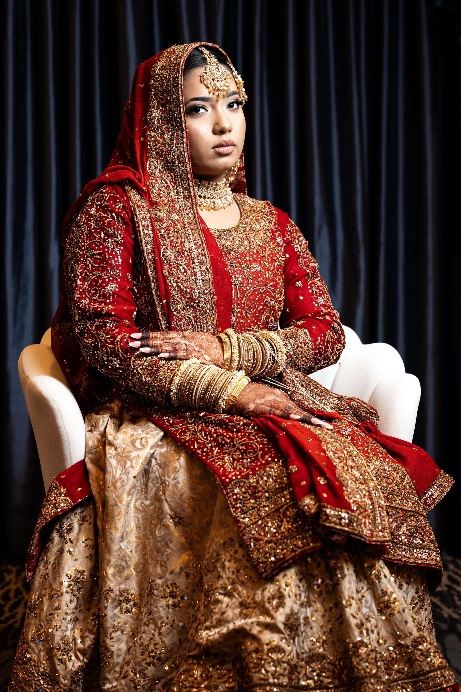 A bride in ornate red and gold Indian bridal attire sits elegantly in a white chair.