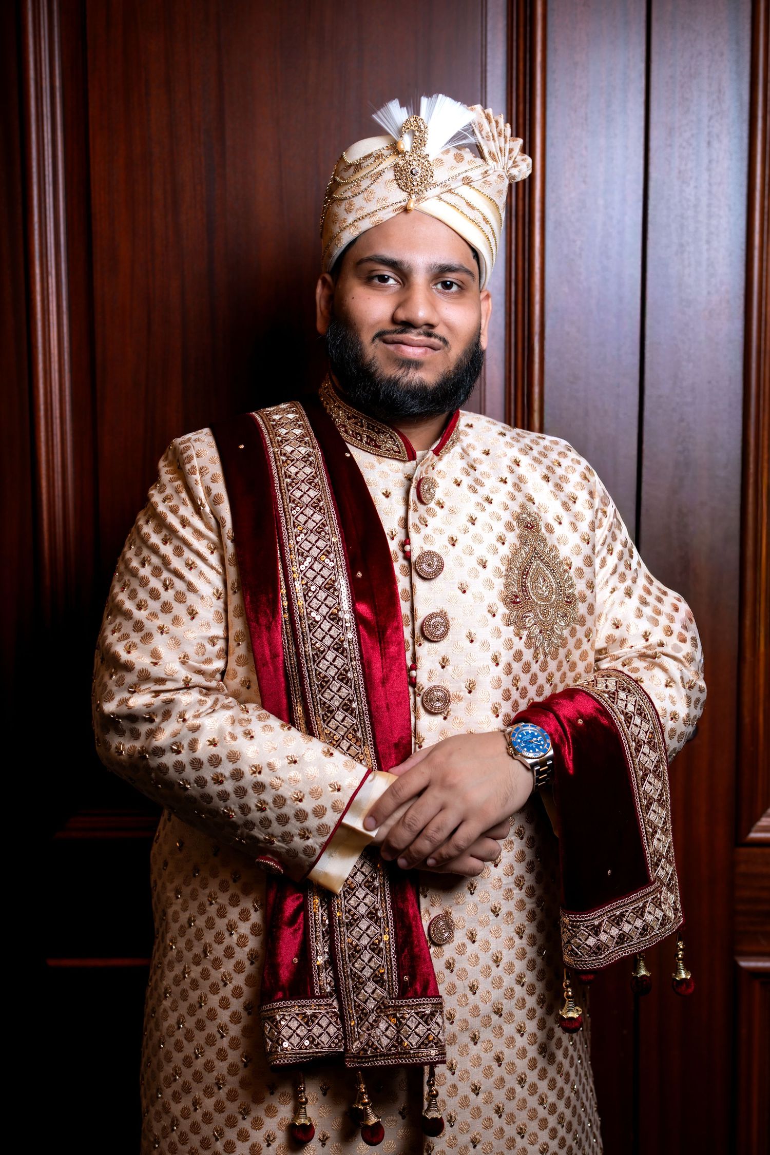 Indian groom in gold sherwani with red velvet dupatta and ornate turban posing elegantly indoors.