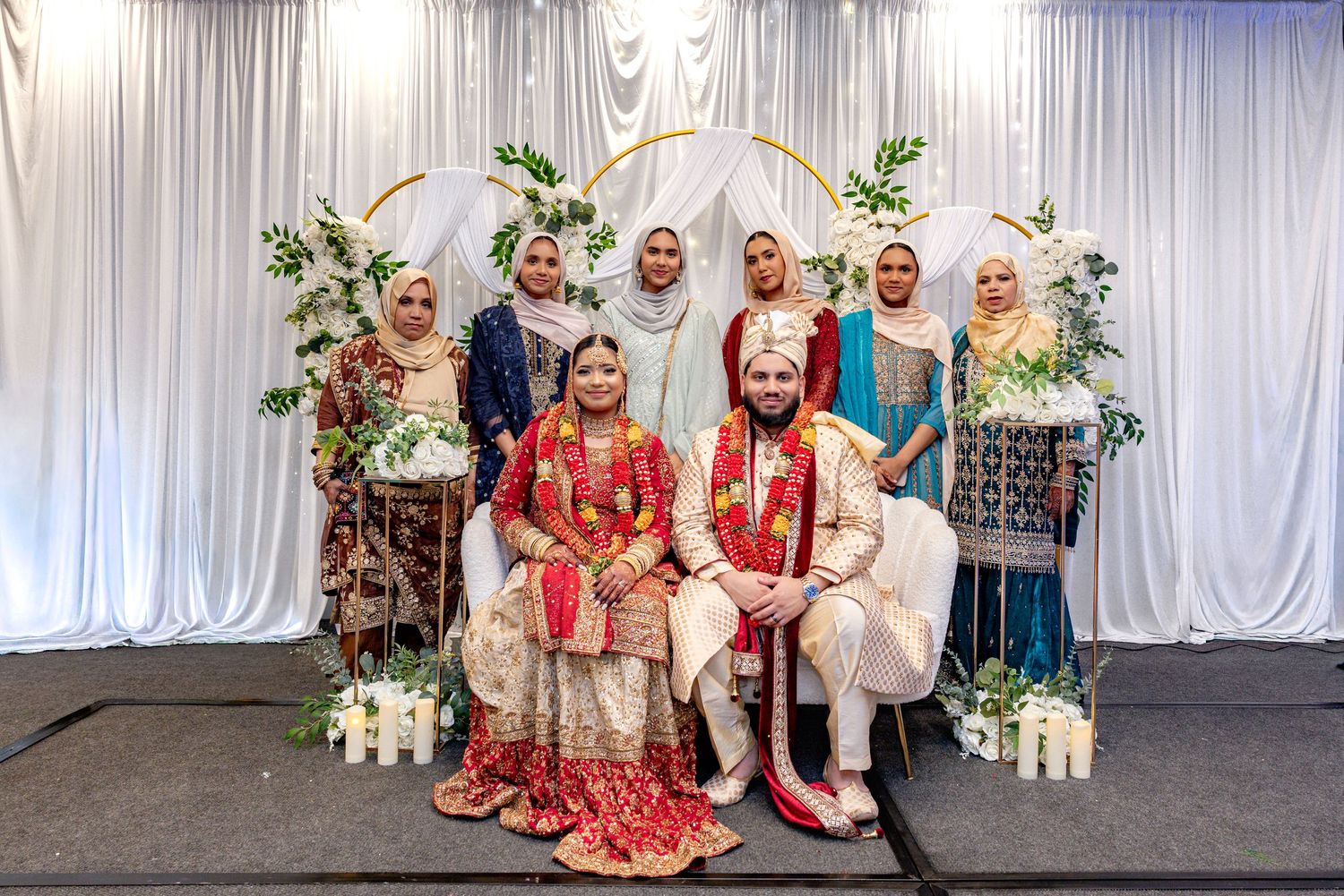 South Asian wedding couple in traditional red and gold attire pose with bridesmaids before floral arch.