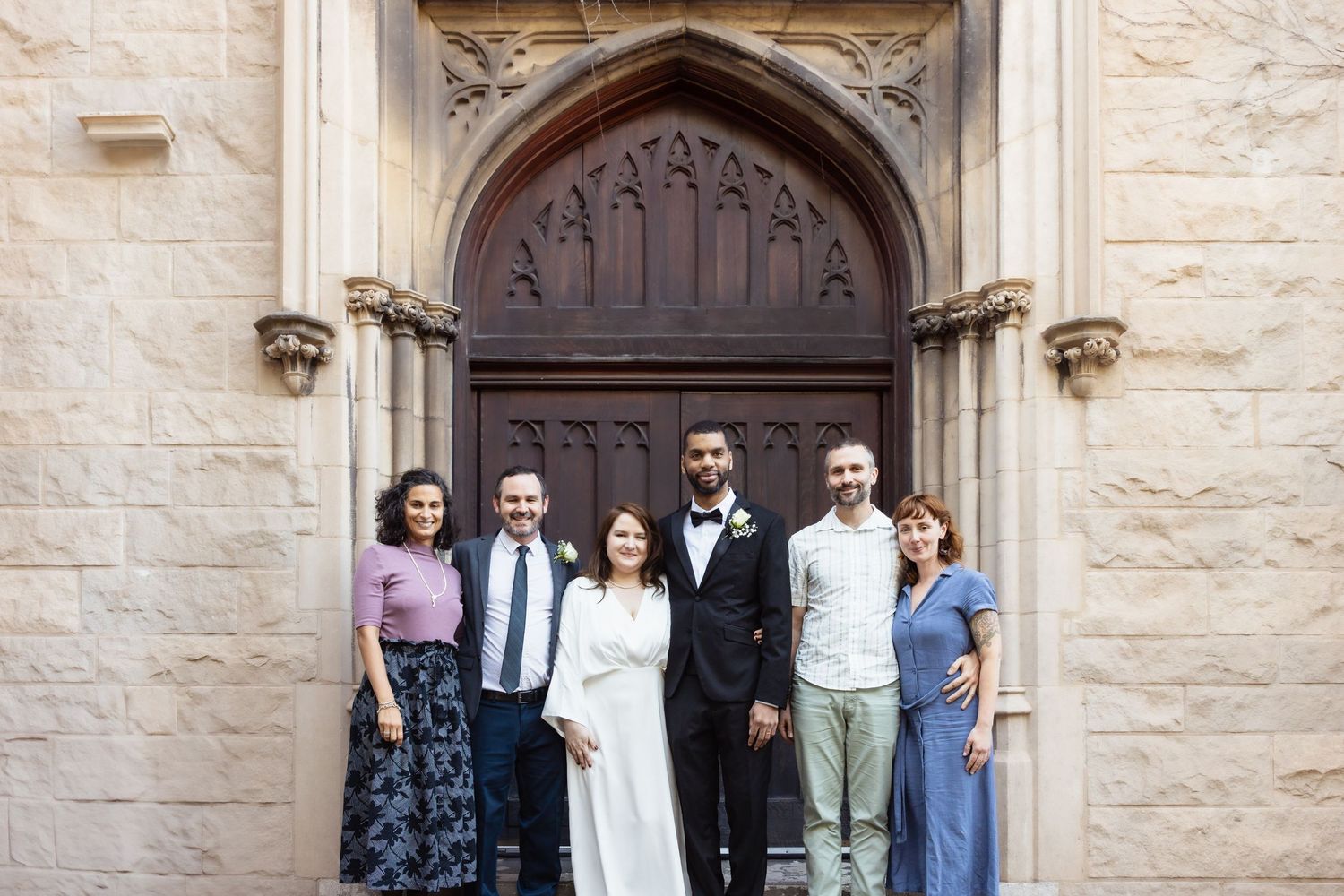 Wedding couple with six guests posing before grand Gothic stone church entrance.