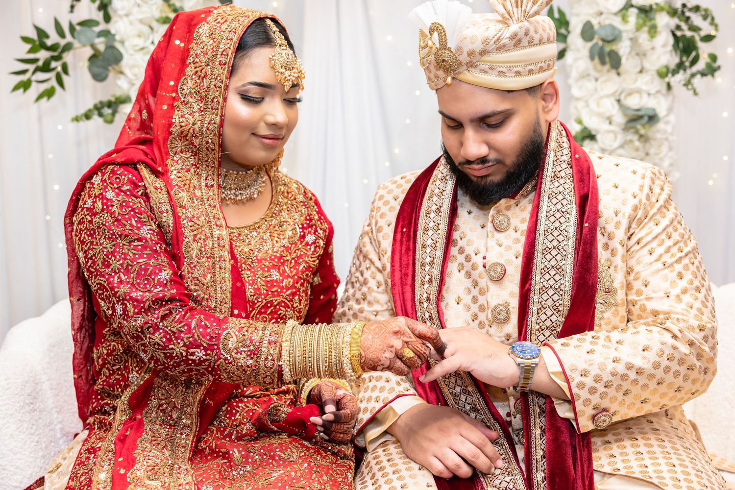 South Asian bride and groom in traditional red and gold wedding attire exchanging rings.