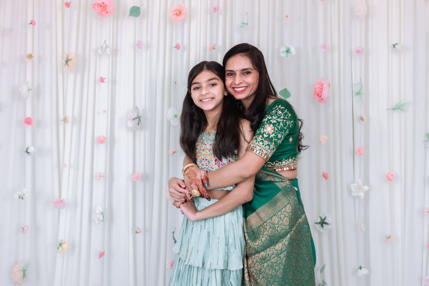 Woman in green saree embraces young girl in mint lehenga at Indian celebration with floral backdrop.