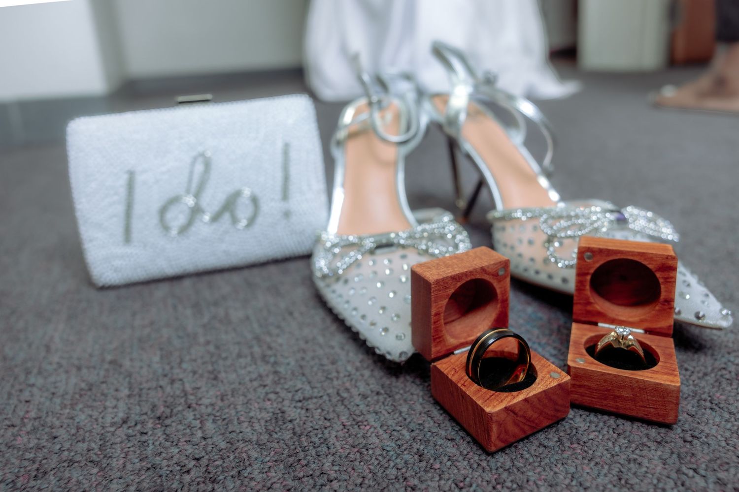 Wedding rings in wooden boxes beside silver glitter heels and an I do clutch purse on gray carpet.