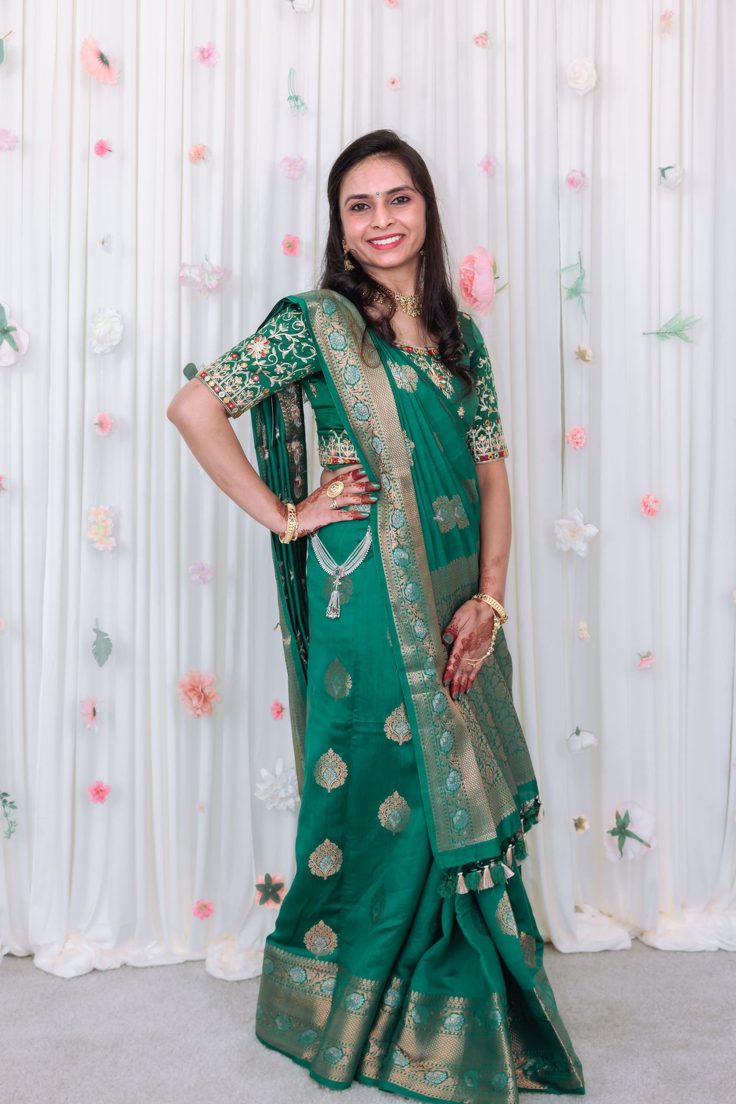 Smiling Indian woman in rich green gold Banarasi saree poses at decorated wedding celebration backdrop.