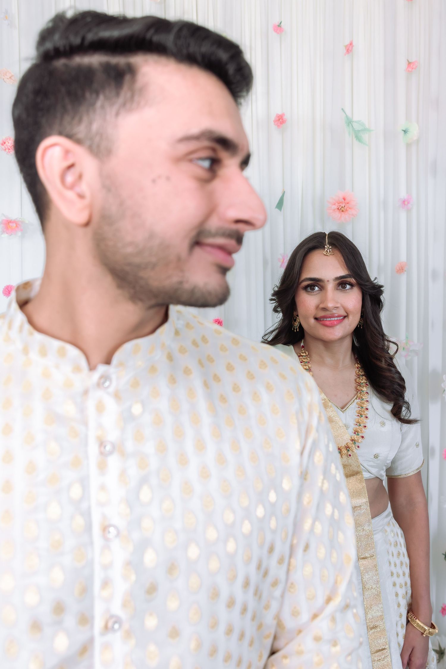 Indian groom in white kurta with bride in white lehenga smiling in background at engagement ceremony.