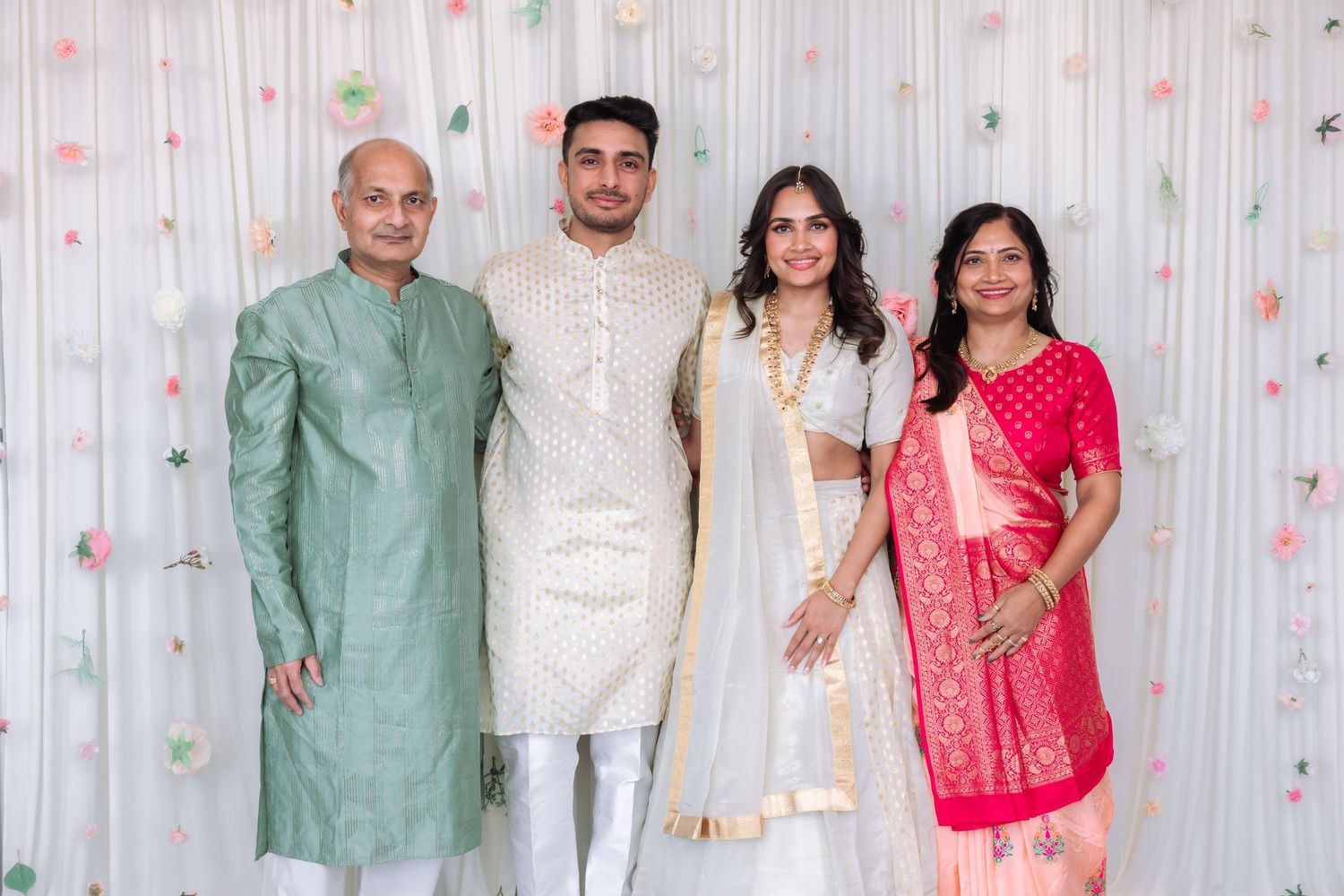 Indian wedding couple in white traditional attire poses with parents wearing pink saree and sage green kurta before floral backdrop.