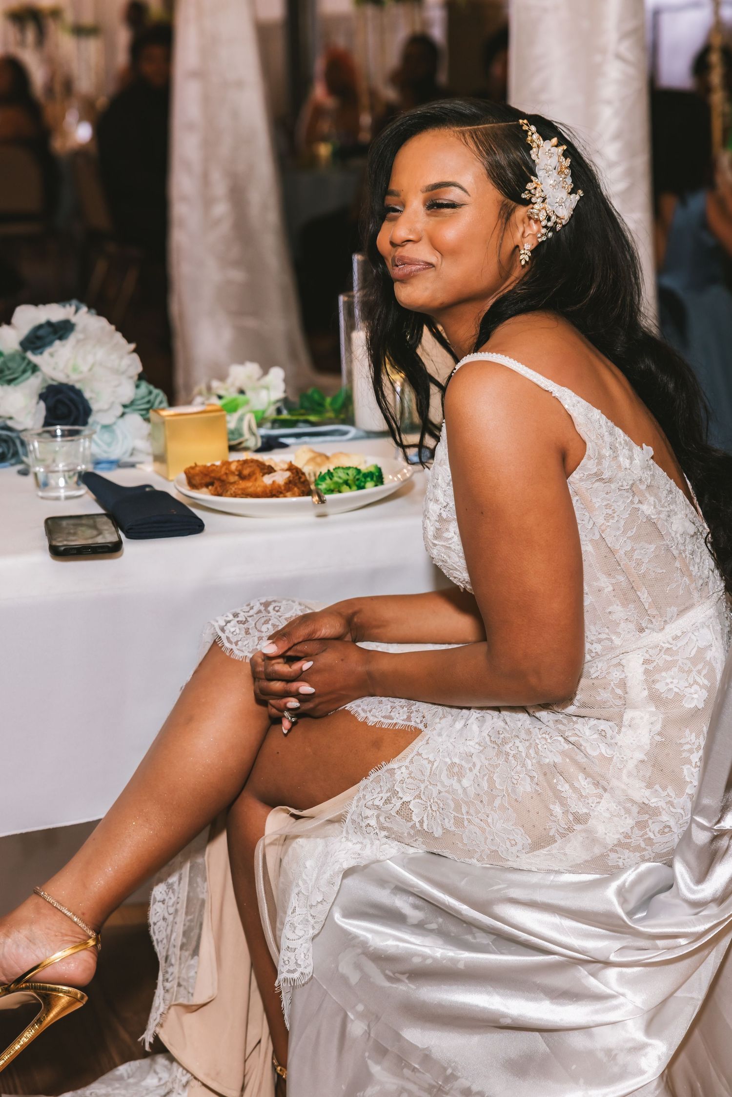 Bride in lace wedding gown sits at reception table smiling with floral hair accessory.