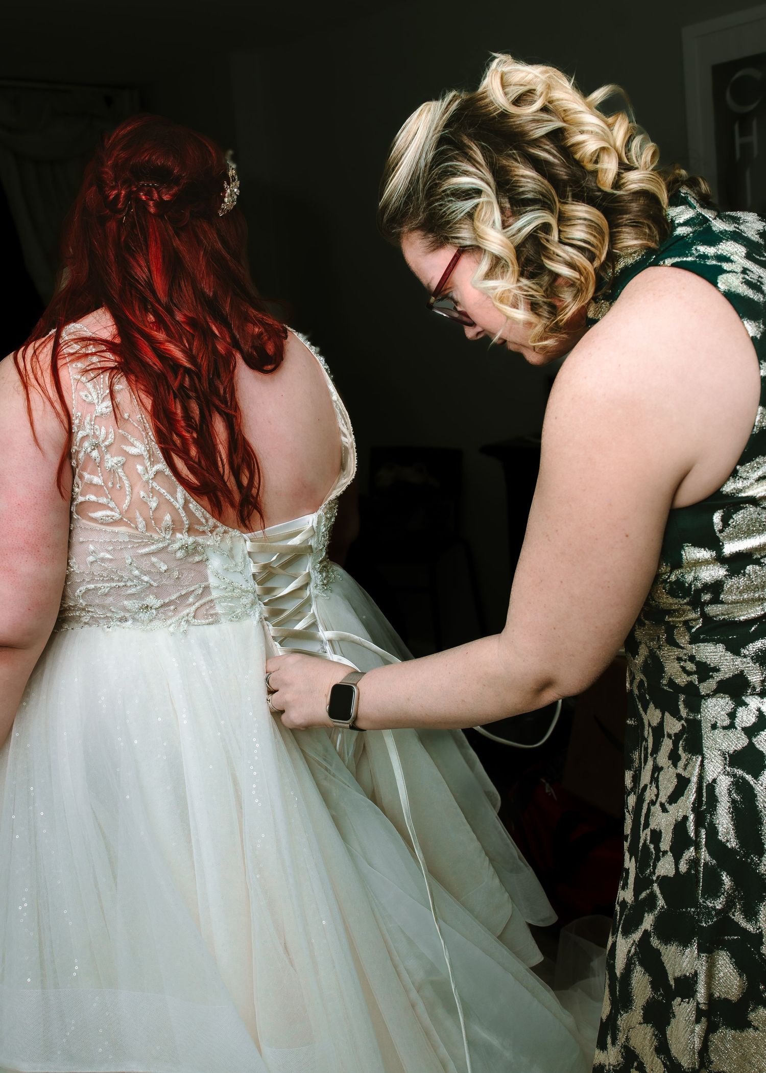 Redhead bride in lace wedding gown being assisted by curly-haired bridesmaid in green dress.