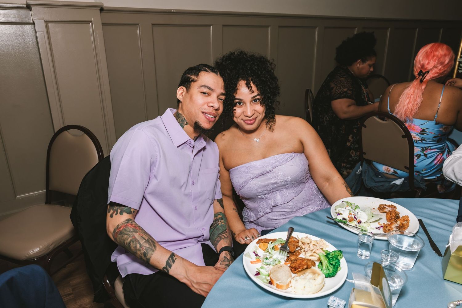 A tattooed man in lavender and a woman in a strapless dress smile together at a wedding reception dinner table.