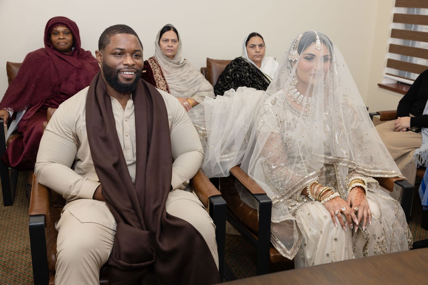 A groom in cream attire and bride in white bridal dress sit together during an Islamic wedding ceremony.