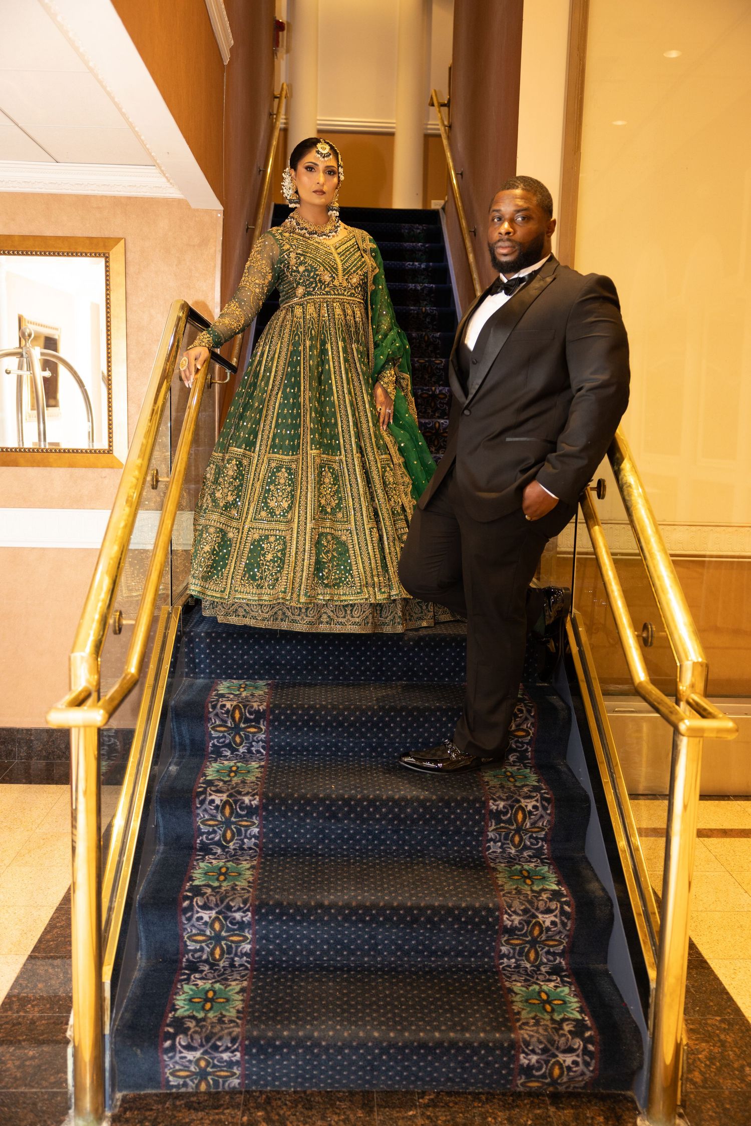 Couple poses on gold-railed staircase, bride in green embroidered lehenga, groom in black tuxedo.