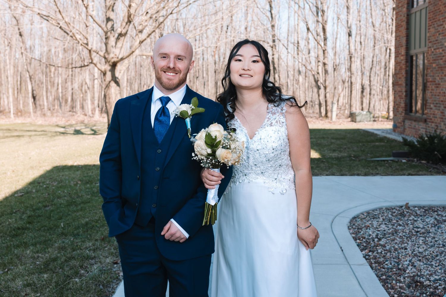 Smiling bride and groom pose outdoors in wedding attire, she holds a white bouquet, bare trees in background.