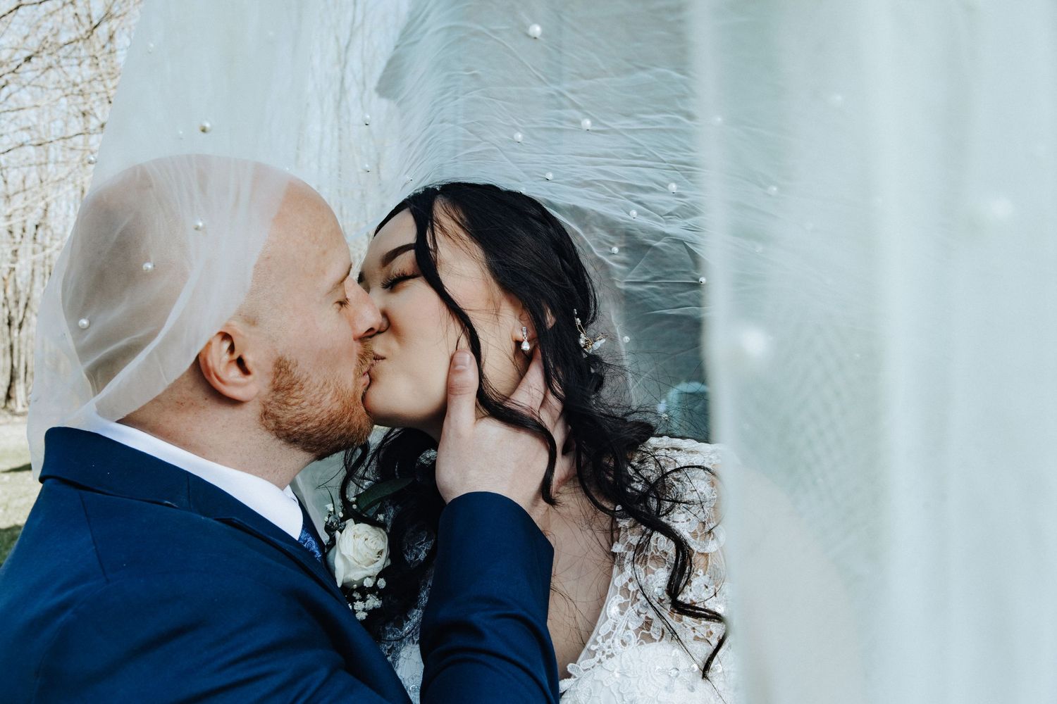 Newlyweds kissing tenderly beneath a flowing bridal veil outdoors on their wedding day.
