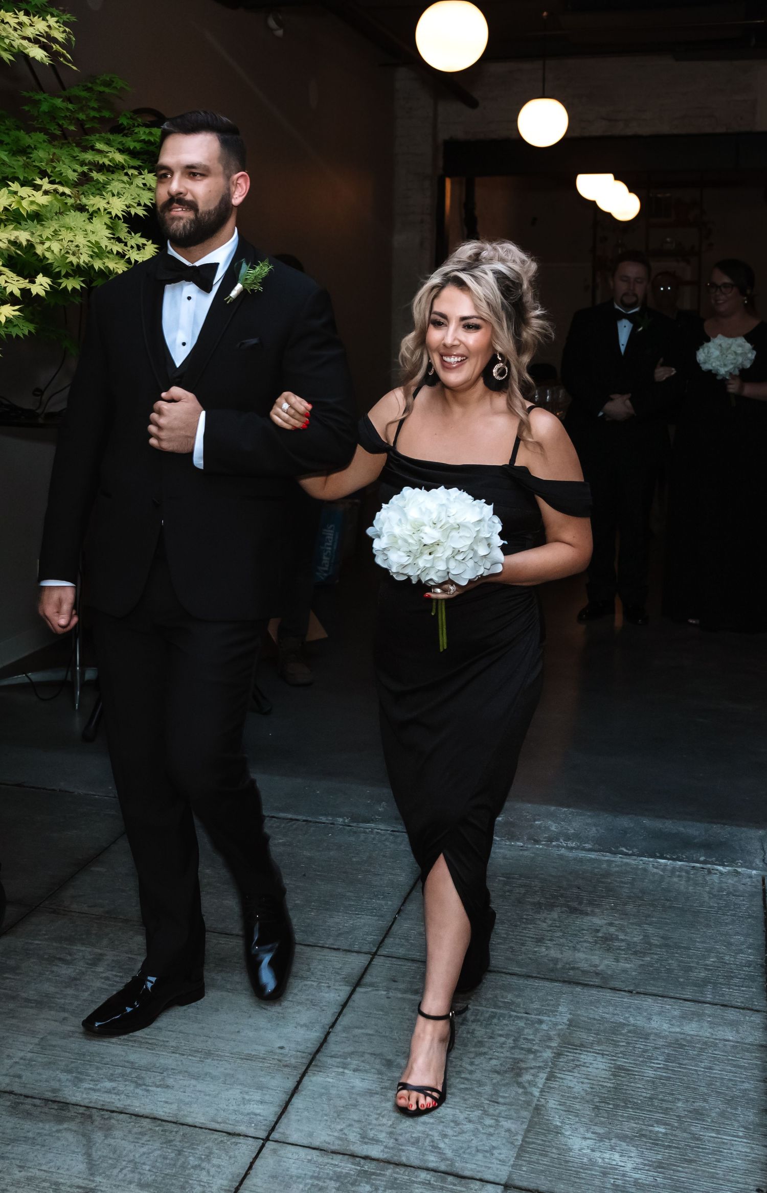 Groomsman in tuxedo escorting bridesmaid in off-shoulder black dress holding white bouquet.