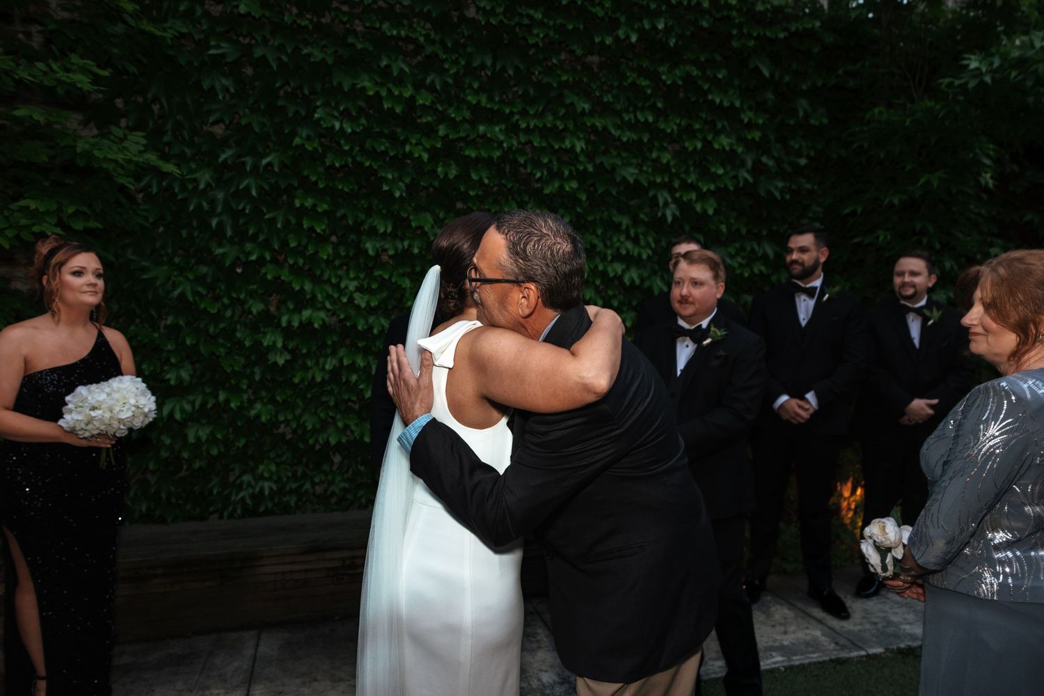 Bride in white gown embracing escort at outdoor wedding reception with guests applauding in background.