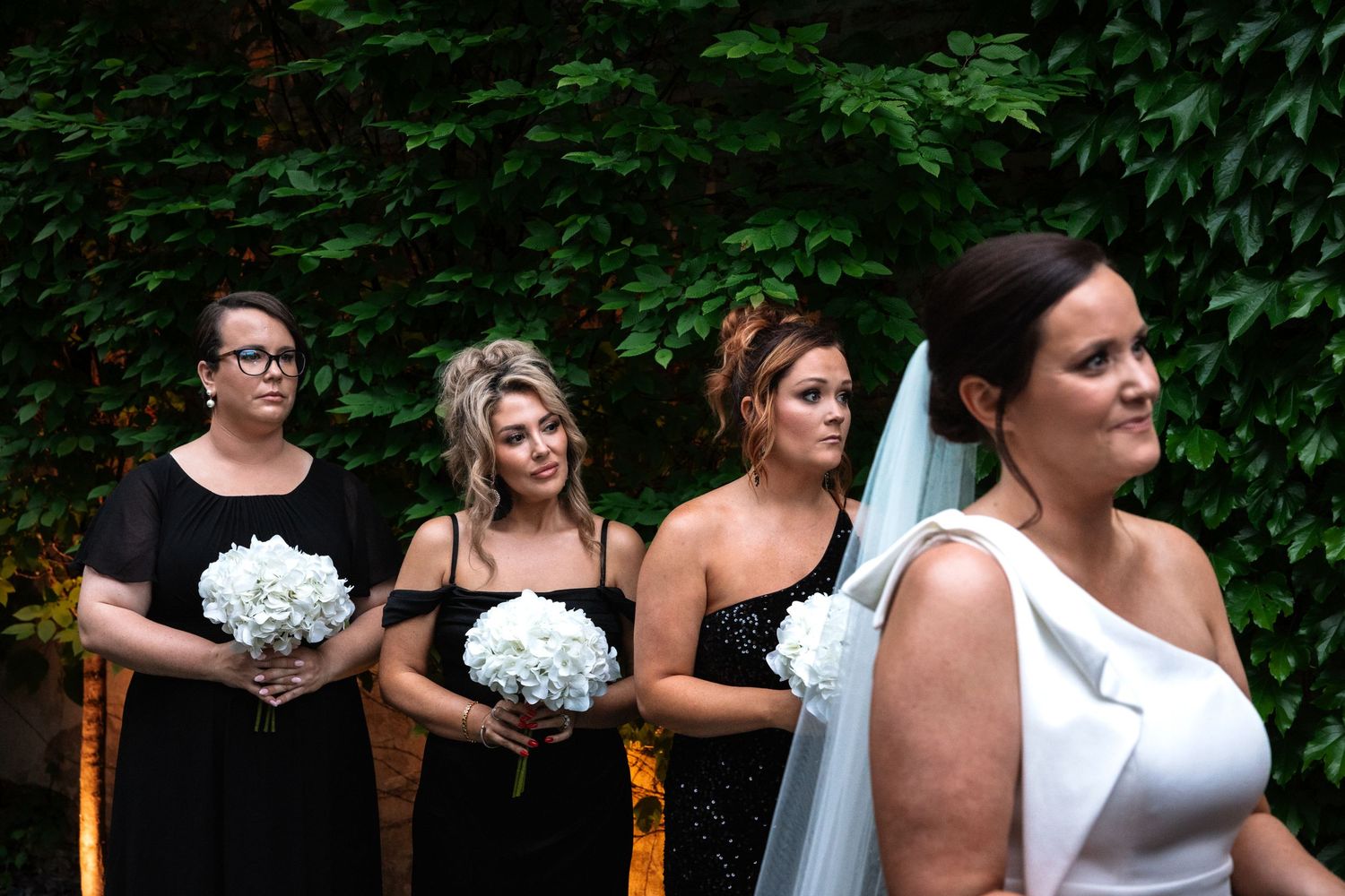 Four bridesmaids in black dresses holding white floral bouquets stand beside bride in white gown outdoors.