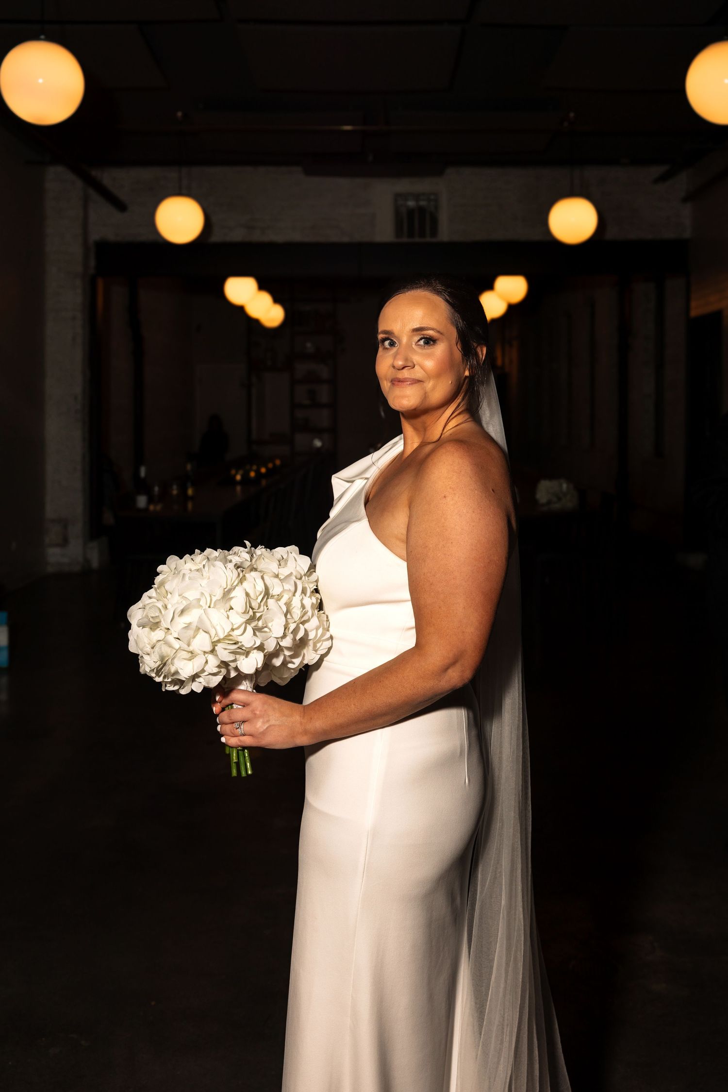 Bride in elegant white satin gown with veil holding white hydrangea bouquet in moody venue.