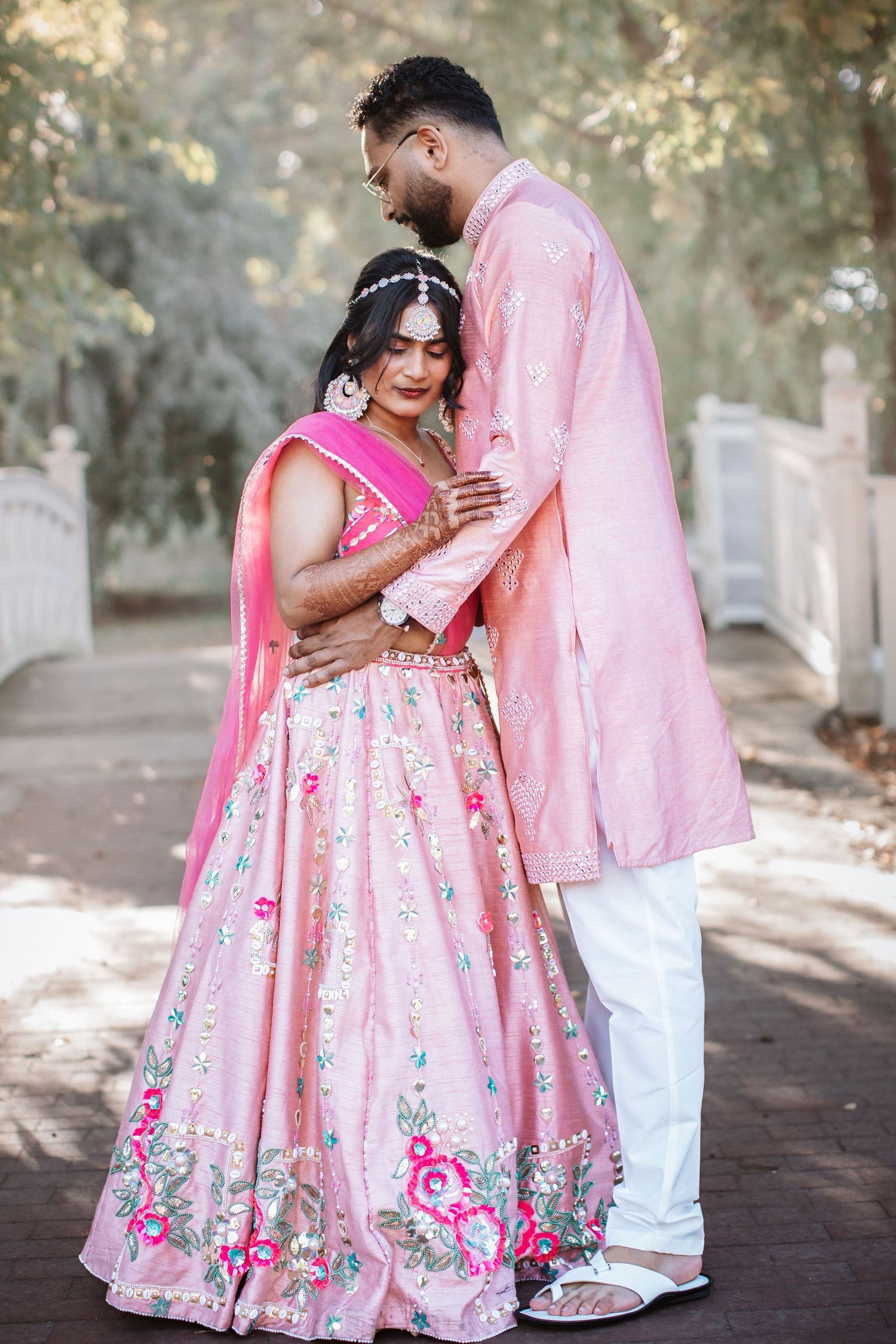 Indian couple in pink traditional attire embracing on a tree-lined garden path.