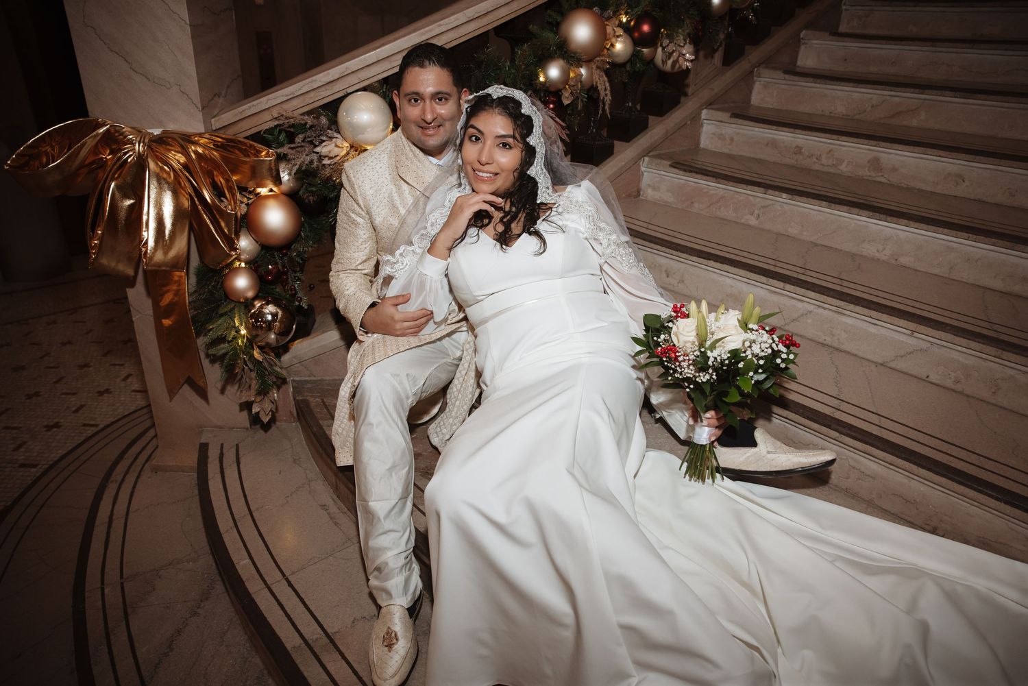 Bride and groom posing on ornate marble staircase decorated with gold Christmas ornaments and garland.