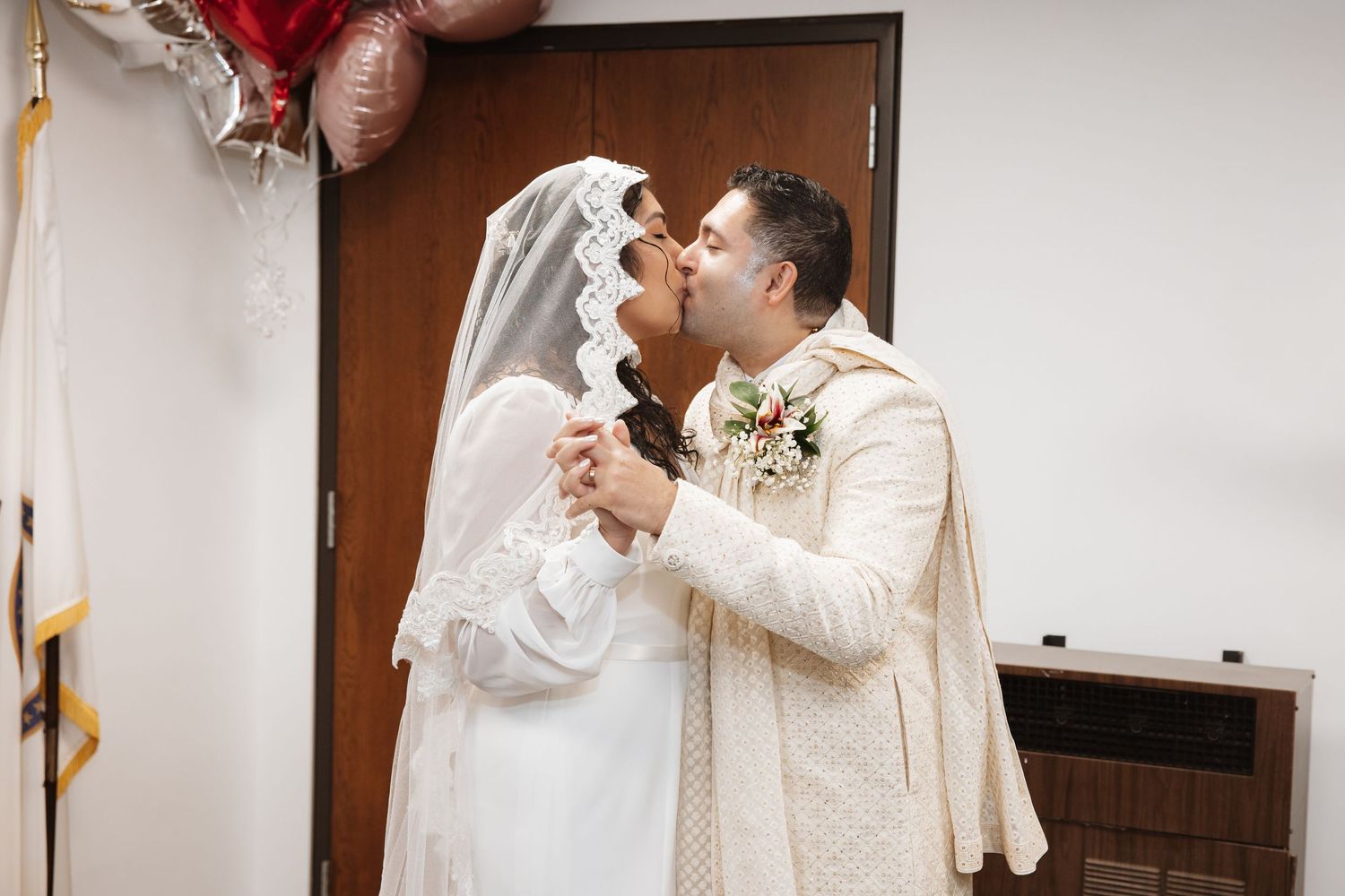 Newlywed couple shares first kiss as bride in white lace veil and groom in ivory sherwani hold hands.
