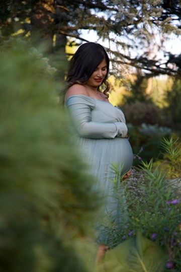 Pregnant woman in sage green off-shoulder gown standing among lush garden foliage at golden hour.